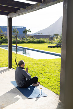 Senior Asian man sitting cross-legged wearing athletic wear on blue mat by lap pool, copy space
