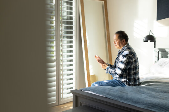 Asian man wearing flannel shirt sitting on bed edge rubbing hands near shutters, copy space