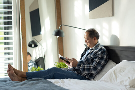 Senior Asian man sitting on bed reading paperback, wearing plaid shirt and jeans by shutters