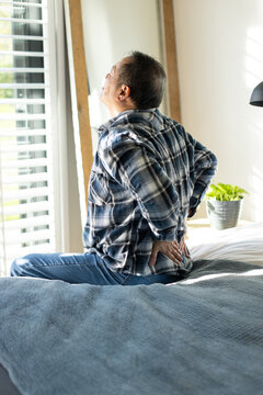 Senior Asian man sitting on edge of bed wearing plaid flannel shirt, jeans, pressing lower back
