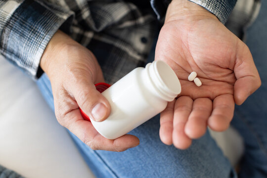Senior Asian in plaid shirt, jeans at home holding red-capped bottle, pouring two pills into palm