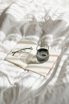 Open book with glasses and water glass on white bed linen