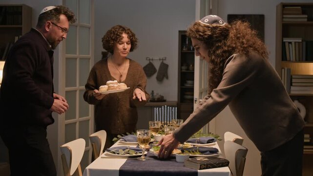 Caucasian Jewish adults serving traditional food and setting dinner table with menorah for Hanukkah. Footage suited for Jewish holiday campaigns, cultural promotions, and festive marketing