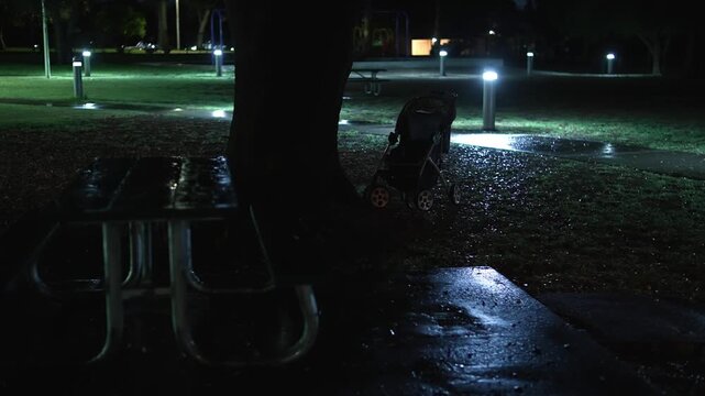 A lonely baby stroller stands beneath a tree in a dark park at night near wet picnic tables and an illuminated pathway, creating an eerie mysterious atmosphere
