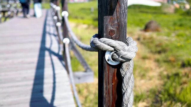 Detailed shot of a thick marine-style rope tied to a wooden pillar with a metal ring, serving as a boundary railing for a pedestrian pathway or bridge outdoors. 