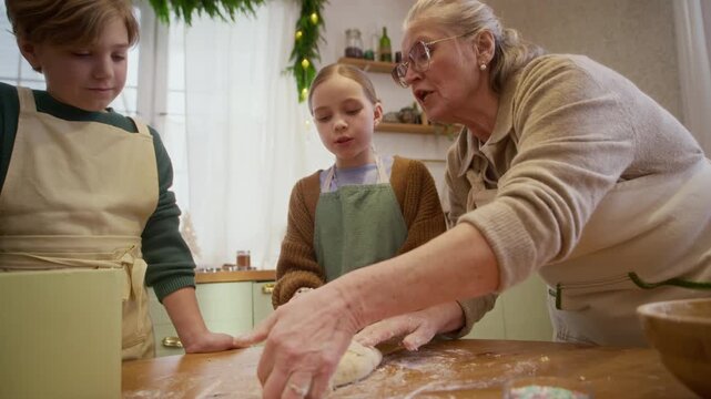 Senior Caucasian grandmother teaching children kneading dough on kitchen table. Clip suited for family lifestyle advertising, holiday culinary campaigns, or baking workshop promos