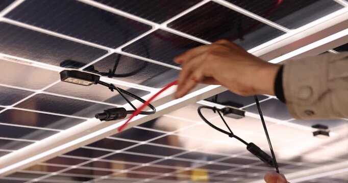 Close-up of a technician&rsquo;s hands routing electrical cables beneath solar panels. Visible connectors, junction boxes, and mounting rails show wiring details during photovoltaic system installation.