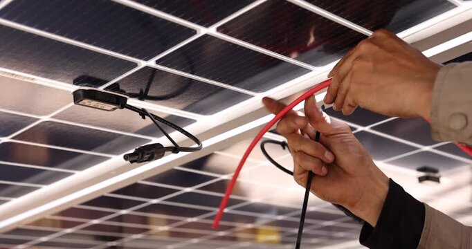 Close-up of a technician&rsquo;s hands routing electrical cables beneath solar panels. Visible connectors, junction boxes, and mounting rails show wiring details during photovoltaic system installation.
