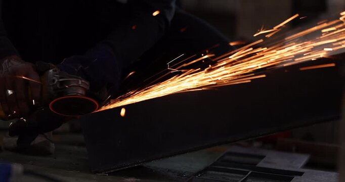 Close-up of a worker using an angle grinder on a steel workpiece in a workshop. Bright orange sparks streak across the dark background during metal grinding and fabrication.