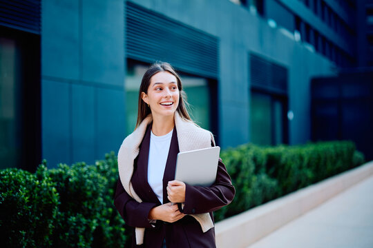 Young woman smiling, carrying a laptop, and walking confidently in a modern city street, reflecting success and a professional career