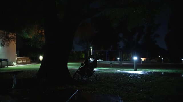 An abandoned baby stroller stands beneath a tree in an empty park at night near a wet pathway and playground, creating a mysterious and unsettling atmosphere