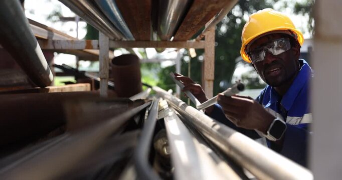 Worker wearing a yellow hard hat, protective goggles, and blue workwear selects steel stock from a storage rack for workshop use. 