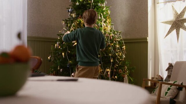 Caucasian boy hanging ornaments on illuminated Christmas tree, viewed from rear. Footage suitable for holiday advertising, winter campaigns, and festive family promos
