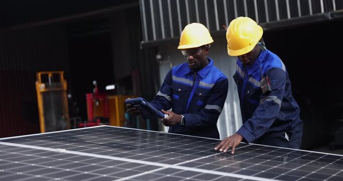Two technicians wearing blue workwear and yellow hard hats inspect solar panels before installation, with one holding a digital tablet.