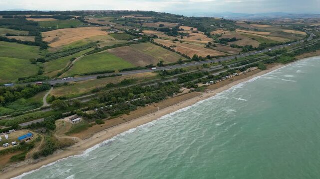Aerial view of the Molise coast in Italy, showing a sandy beach along the turquoise Adriatic Sea. A coastal road, railway, highway, and seaside campsite border rolling agricultural fields and