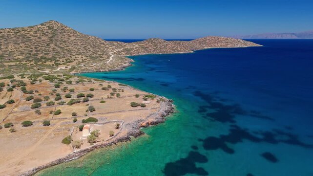 Drone pulling back from a small coastal church on the shore of Elounda, revealing the clear sea and dry Mediterranean landscape