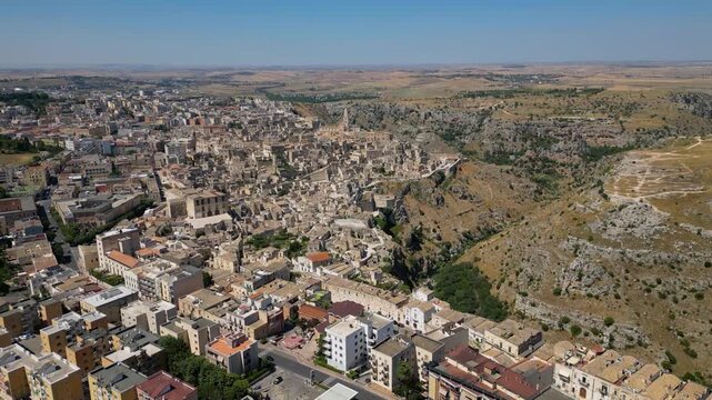 A high aerial panorama of Matera, Italy, showing the famous ancient Sassi quarter built along the edge of a dramatic canyon. The dense stone architecture, cathedral tower, and surrounding rugged