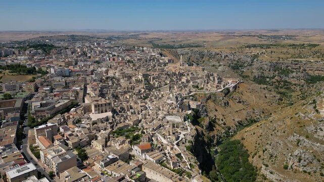 A high aerial panorama of Matera, Italy, showing the famous ancient Sassi quarter built along the edge of a dramatic canyon. The dense stone architecture, cathedral tower, and surrounding rugged