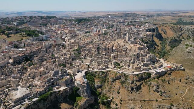 A high aerial panorama of Matera, Italy, showing the famous ancient Sassi quarter built along the edge of a dramatic canyon. The dense stone architecture, cathedral tower, and surrounding rugged