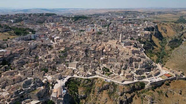 A high aerial panorama of Matera, Italy, showing the famous ancient Sassi quarter built along the edge of a dramatic canyon. The dense stone architecture, cathedral tower, and surrounding rugged