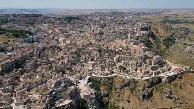 A high aerial panorama of Matera, Italy, showing the famous ancient Sassi quarter built along the edge of a dramatic canyon. The dense stone architecture, cathedral tower, and surrounding rugged