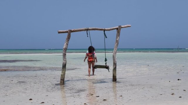 Woman sitting on a wooden swing suspended over shallow turquoise sea water at Karimunjawa island, tropical vacation lifestyle, leisure travel, summer holiday relaxation, serenity and peace