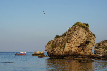 Scenic view of a rugged rock formation Isola Bella in the sea with a distant boat and flying bird, perfect for travel concepts and marine themes © Olga Yashnikova