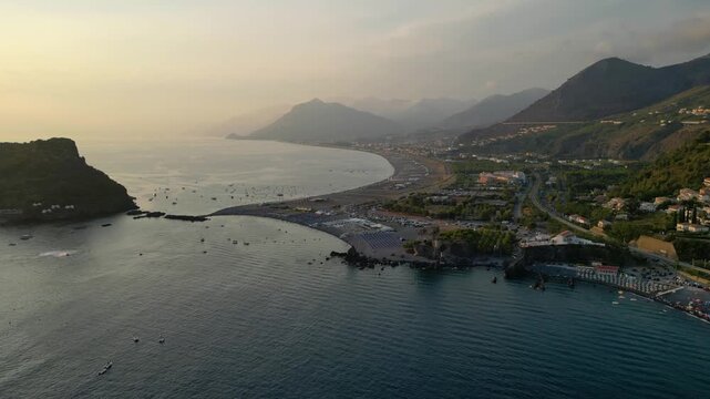A breathtaking sunset aerial view captures Isola del Dino and the bay at Praia a Mare in Calabria, Italy. Boats navigate the deep blue waters along a sprawling beach, framed by dramatic coastal