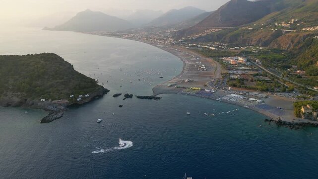 A breathtaking sunset aerial view captures Isola del Dino and the bay at Praia a Mare in Calabria, Italy. Boats navigate the deep blue waters along a sprawling beach, framed by dramatic coastal