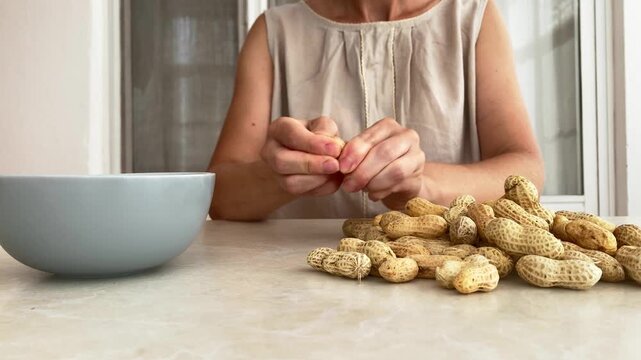 A woman is sitting at a table and peeling fresh peanuts, a natural product of plant origin, a source of plant protein. The topic is healthy eating and nutritious snacks