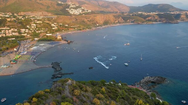 A breathtaking sunset aerial view captures Isola del Dino and the bay at Praia a Mare in Calabria, Italy. Boats navigate the deep blue waters along a sprawling beach, framed by dramatic coastal