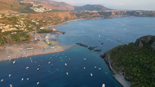 A breathtaking sunset aerial view captures Isola del Dino and the bay at Praia a Mare in Calabria, Italy. Boats navigate the deep blue waters along a sprawling beach, framed by dramatic coastal