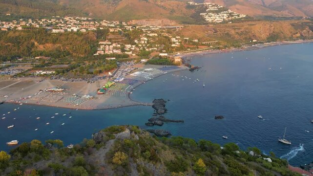 A breathtaking sunset aerial view captures Isola del Dino and the bay at Praia a Mare in Calabria, Italy. Boats navigate the deep blue waters along a sprawling beach, framed by dramatic coastal