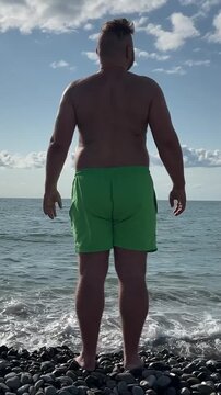 Vertical travel video. 60 fps. Back view. Young adult tanned fat white man with big belly, green shorts on sea pebble beach looks around at summer vacation at sun day. Blue sky. Ocean waves. Tourism