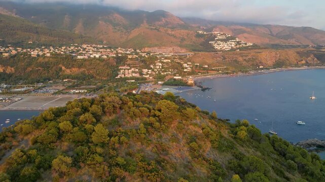 A breathtaking sunset aerial view captures Isola del Dino and the bay at Praia a Mare in Calabria, Italy. Boats navigate the deep blue waters along a sprawling beach, framed by dramatic coastal