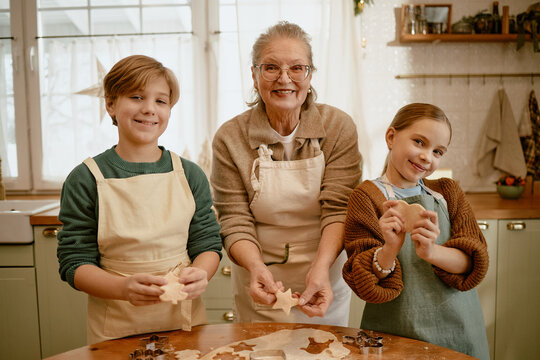 Portrait of senior Caucasian woman, male teenager and child girl baking cookies together in kitchen, smiling at camera. Image suitable for family lifestyle advertising and holiday baking promotions