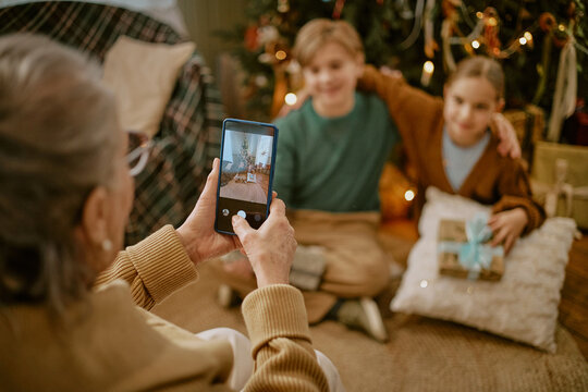 Senior Caucasian woman taking smartphone photo of grandchildren celebrating Christmas. Ideal for holiday marketing, mobile technology promotion and family campaigns