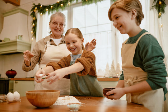 Senior Caucasian grandmother baking pastry with grandchildren in kitchen, breaking eggs into wooden bowl. Concept represents family bonding, holiday cooking, culinary advertising campaigns
