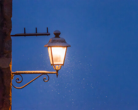 View of a glowing vintage street lamp covered in light snow against a dark blue evening sky in Castelluccio, Umbria, Italy.