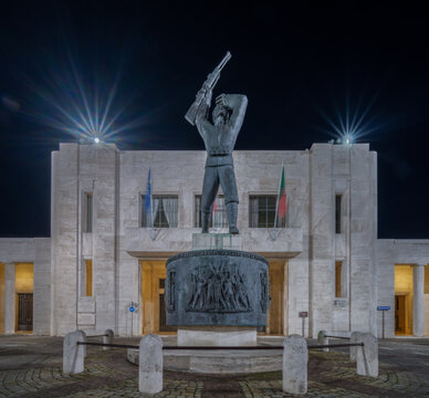 View of the Monumento ai Caduti, a bronze soldier statue with a rifle standing on a stone base in front of an illuminated building at night in Corridonia, Marche, Italy.