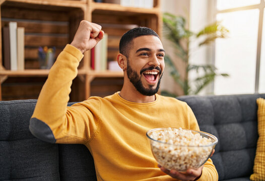 Excitement young man cheering with fist pump sitting on gray sofa holding large bowl of popcorn in sunny living room