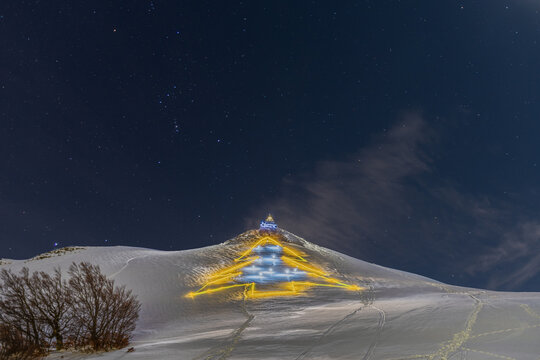 View of a large illuminated Christmas tree shape created with lights on a snow-covered mountain slope under a starry night sky in Bolognola, Marche, Italy.