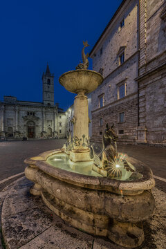 View of the historic fountain in Piazza del Popolo with illuminated stone architecture and a clear evening sky, Ascoli Piceno, Marche, Italy.