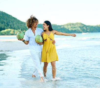 A young couple laugh and dance along a tropical beach, the man holding a fresh coconut as waves lap the shoreline. The scene captures romantic summer vacation vibes, carefree joy and togetherness amid