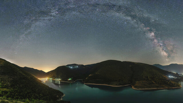 View of Lake Fiastra under a starry night sky with the Milky Way galaxy arching over the dark mountains in Fiastra, Marche, Italy.