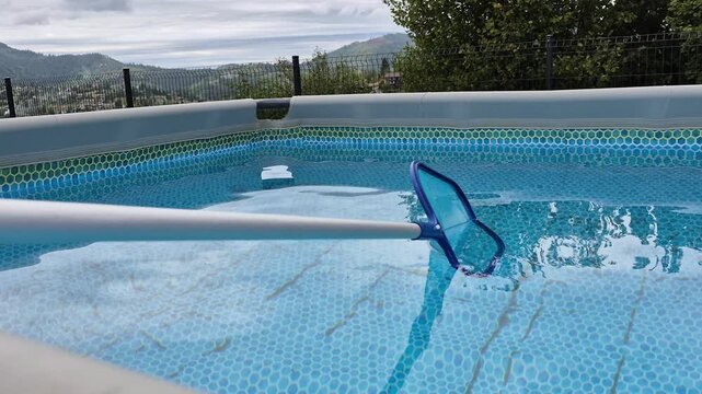 ​Cleaning blue water in an above-ground swimming pool using a skimmer net with beautiful mountain view in the background during summer pool maintenance.