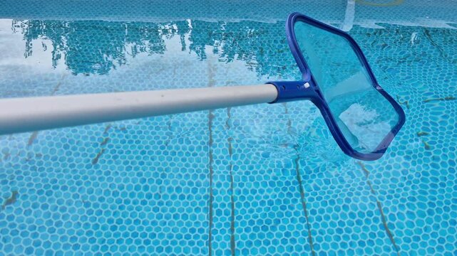 ​Cleaning blue water in an above-ground swimming pool using a skimmer net on a telescopic pole during regular backyard pool maintenance in summer.