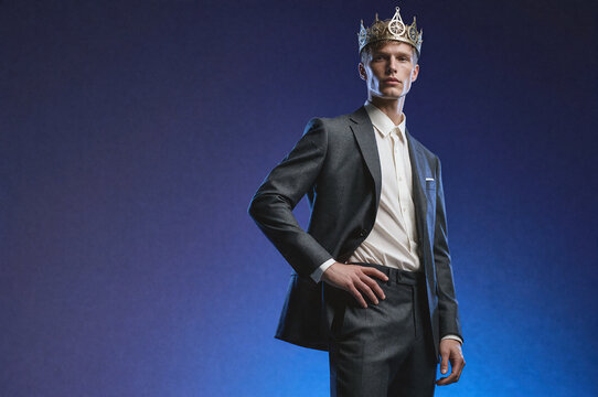Man wearing an intricate gold crown and charcoal suit stands against a blue backdrop. Hard side lighting accentuates the sharp tailoring of his clothing and facial structure.