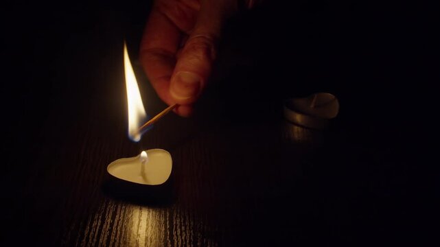 A man's hand lighting heart-shaped candles in a dark room with a burning match