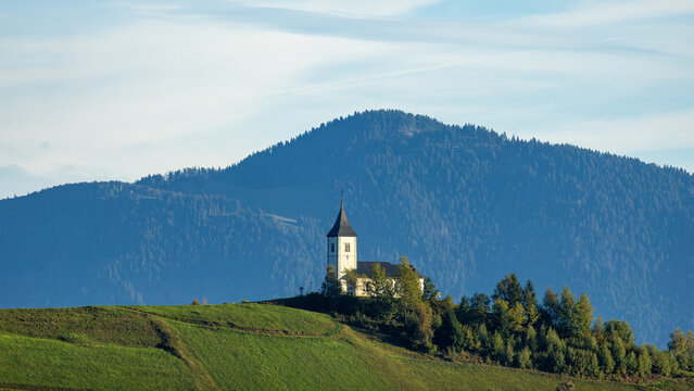 View of the Church of Saints Primus and Felician perched on a grassy hill surrounded by dense forests and rolling mountains in Jamnik, Kranj, Slovenia.